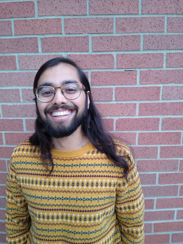 Sumeet in a yellow sweater in front of a brick wall, smiling. 
